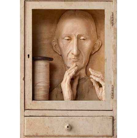 Wooden sculpture of a cabinet with a door and a drawer. Inside the window of the door, there is a bald person with his hands near his face and a large spool of thread to the left of the bust