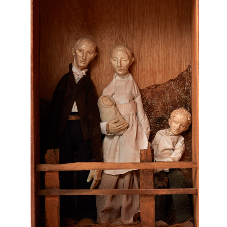 Wooden diorama-style sculpture of 2 adults, one holding a baby, and a child posed behind a fence. They appear to be dressed in 18th or 19th century garb.