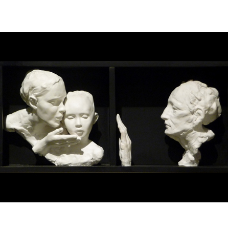 White relief-style sculpture of an adult kissing an adolescent child-- both in profile facing stage right; another profile looks toward them holding out a hand as if to touch, but cannot-- there appears to be a barrier.