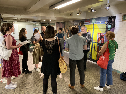 Ron Jenkins reads to a group of ten people in front of a mosaic mural in the Times Square station.